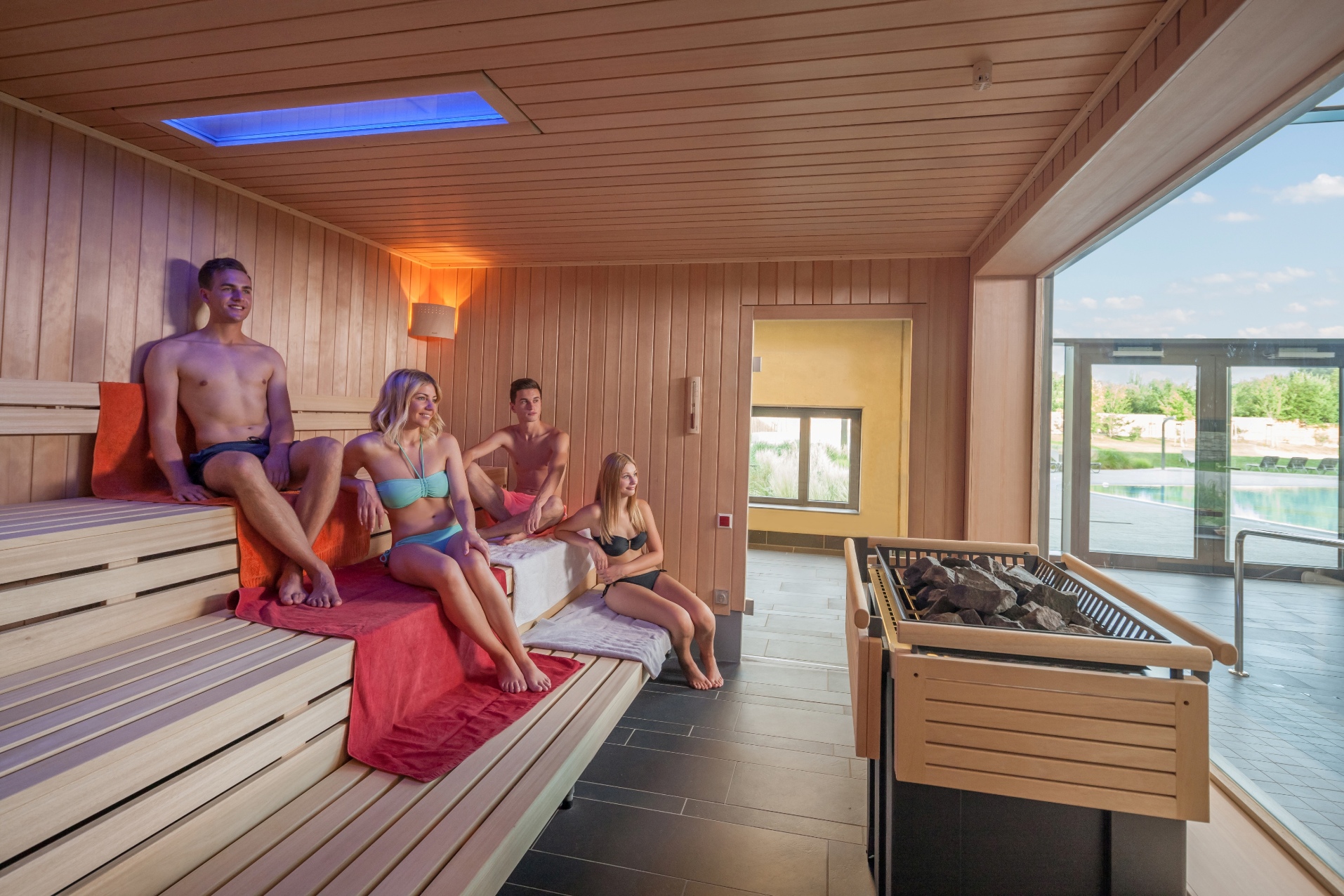Textilsauna in der Wasserwelt mit vier entspannten Gästen auf Holzsaunabänken und Blick durch große Fenster auf den Außenpoolbereich